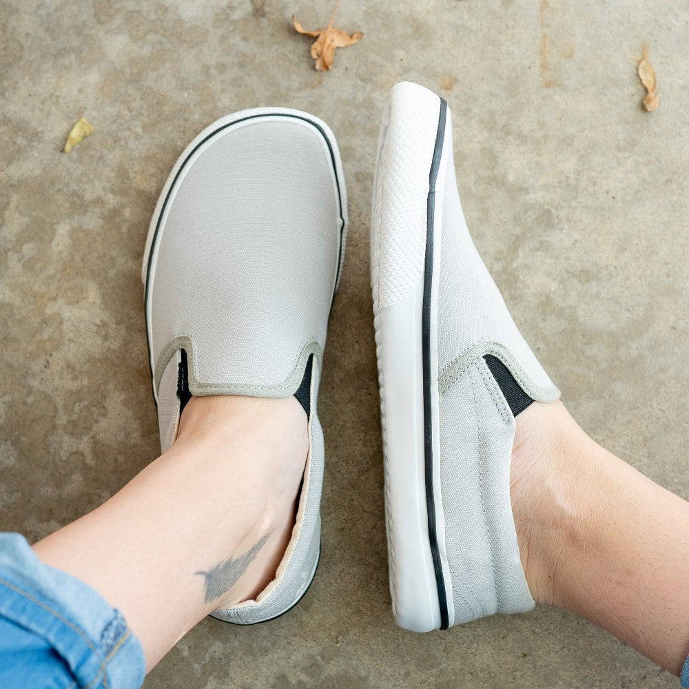 A photo of Bohempia Velik slip on sneakers made from canvas and rubber soles. The sneakers are light grey with white soles with a black stripe around the sole. The left shoe is shown from above and the right shoe is shown from the left side on a woman's feet, with a view of her shins down, on a cement floor. #color_light-grey-white