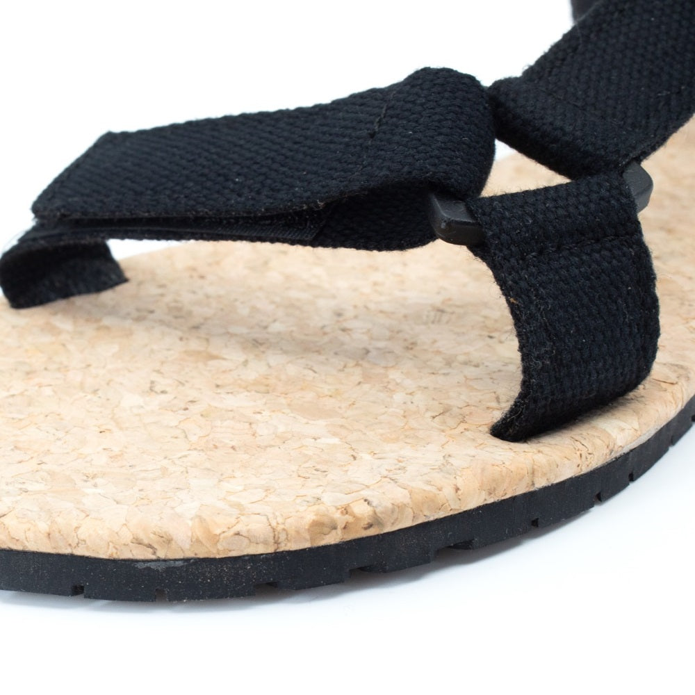 Black Mukishoes Solstice. Sandals have thicker cotton adjustable straps going over the toes and surrounding the ankle with straps connecting the toe and ankle straps. Footbed is quark and sole is a thin black rubber. Left shoe toe strap is shown here against a white background. #color_black