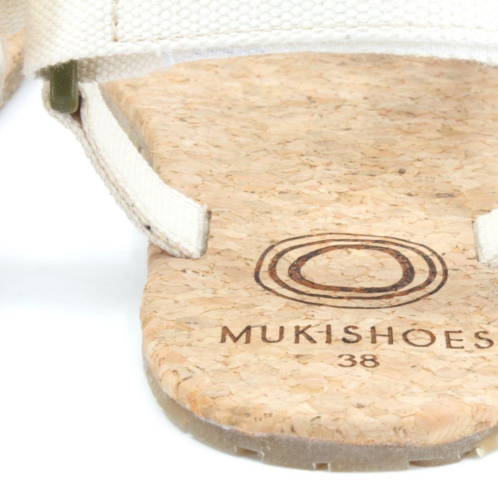 Cream Mukishoes Solstice. Sandals have thicker cotton adjustable straps going over the toes and surrounding the ankle with straps connecting the toe and ankle straps. Footbed is quark and sole is a thin black rubber. Right shoe footbed and strap is shown here from the back against a white background. #color_natural-cream
