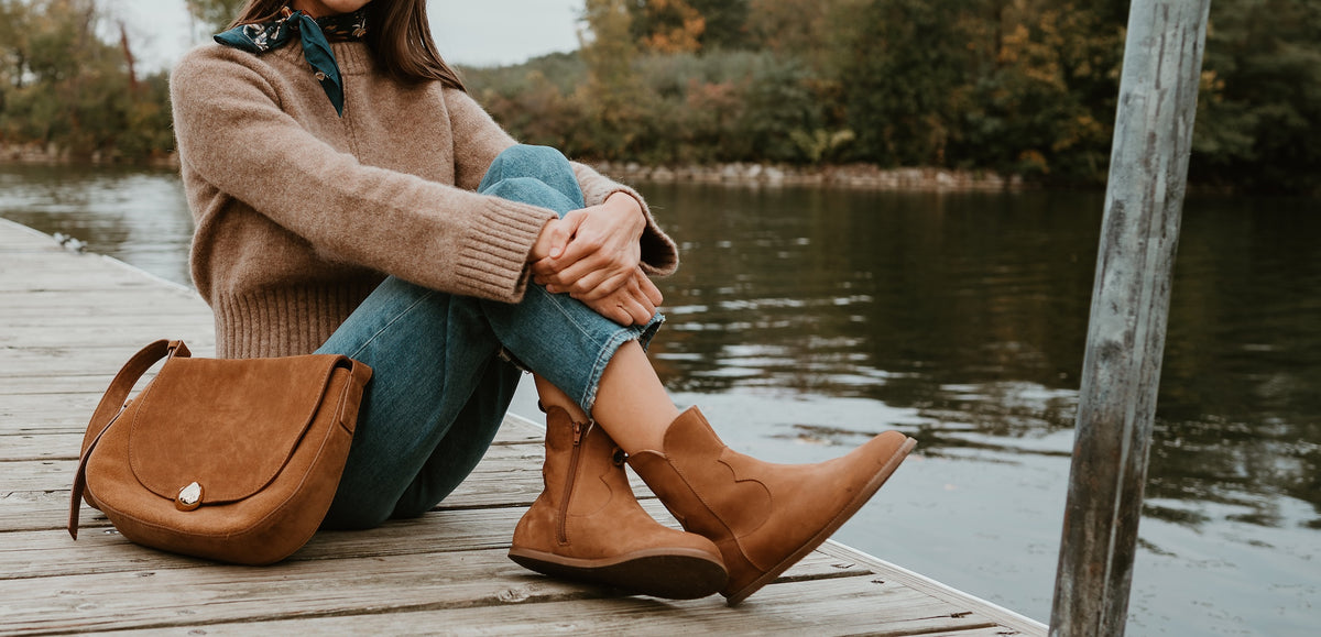 Light brown nubuck leather western Faye boots By Anya with a western shape on both sides of the ankles, a zipper, tan rubber soles, a welt stitching design around the top of the sole, and a pull tab at the top back of the opening. Both boots are shown from the right pointing upwards on a light-skinned woman with long, brown hair sitting on a dock with her feet crossed. #color_cognac