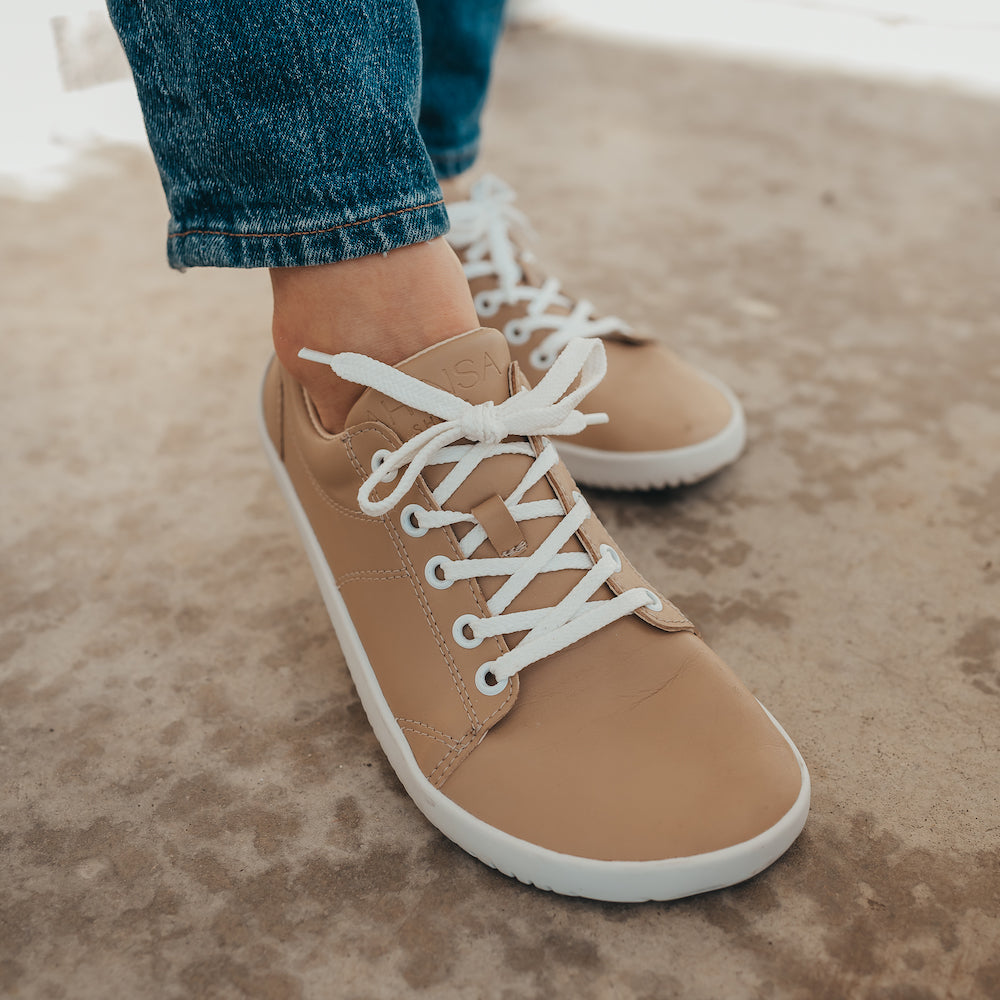 Beige colored vegan sneakers shown on a model. Feet only with one foot slightly in front of the other. Featuring white laces and a white sole.  #color_beige