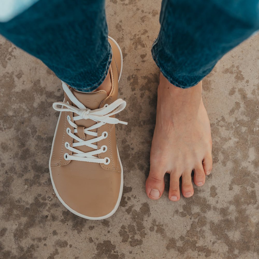 Beige colored vegan sneakers shown on a model. Feet only, top down view of one shoe and one foot that is barefoot.  Featuring white laces and a white sole. #color_beige