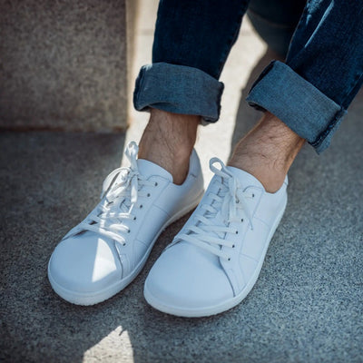 White Angles Fashion Linos leather sneakers. Sneakers are all white and made of Italian leather with rubber soles. Shoes are ankle height and have subtle sneaker stitching and detailing. Both shoes are shown here facing diagonally left on a man wearing rolled-up, dark-wash jeans sitting on back and white speckled pavement next to the same kind of pavement wall. #color_white