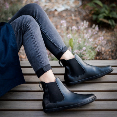 Black smooth leather Angles Nyx Chelsea boot. Boots are simple in design with a long pull tab attached to the heel. Both boots are shown staggered here facing right on a woman with light skin wearing skinny, rolled, washed black jeans sitting on a wooden bench with wild flowers in the background. #color_black