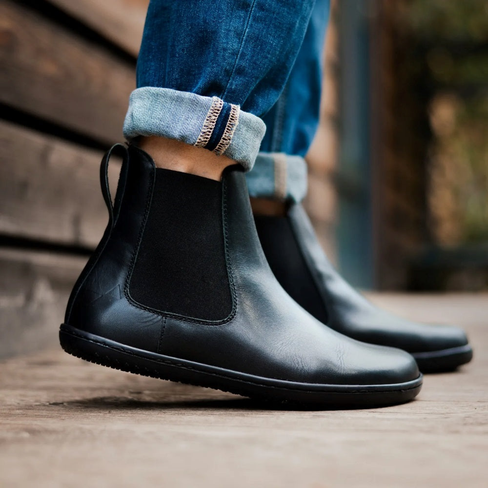 Black smooth leather Angles Nyx Chelsea boot. Boots are simple in design with a long pull tab attached to the heel. Both boots are shown facing right on a person wearing, skinny, rolled-up, medium wash blue jeans standing on a wooden path. #color_black