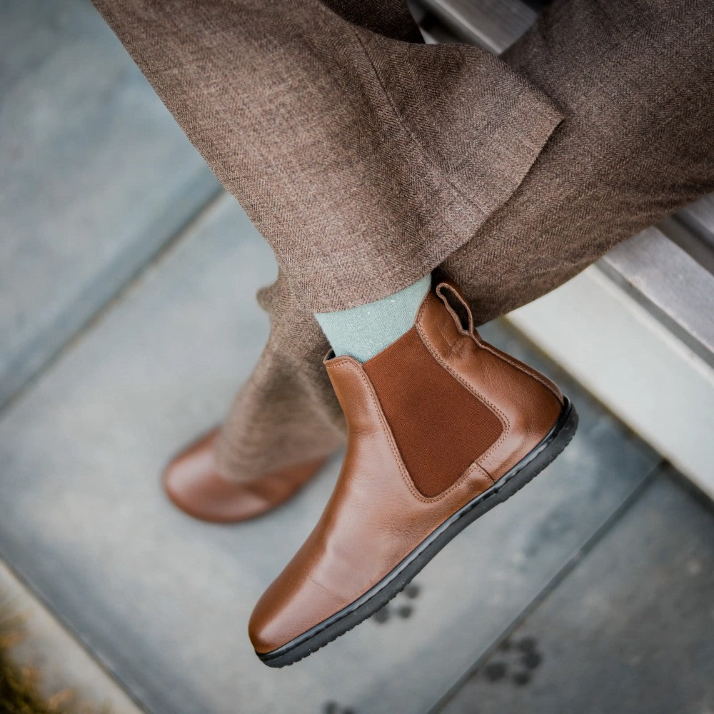 Brown smooth leather Angles Nyx Chelsea boot. Boots are simple in design with a long pull tab attached to the heel. Right boot is in the foreground on a person wearing loose, brown textured slacks and grey socks sitting on a bench with their right ankle crossed over on the left knee. #color_brown