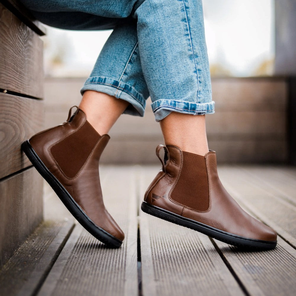 Brown smooth leather Angles Nyx Chelsea boot. Boots are simple in design with a long pull tab attached to the heel. Both boots are shown staggered facing right on a person wearing rolled-up, light-wash, blue jeans sitting on a wooden bench on a wooden path. #color_nut