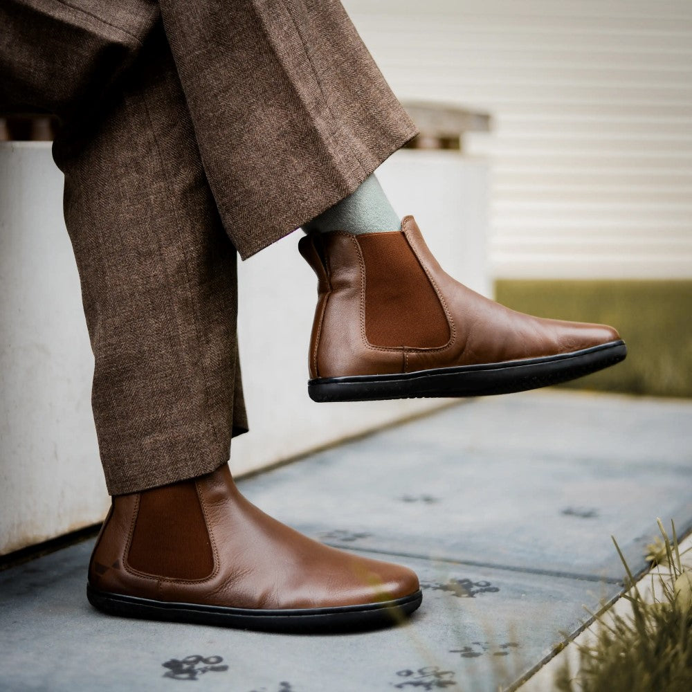 Brown smooth leather Angles Nyx Chelsea boot. Boots are simple in design with a long pull tab attached to the heel. Both boots are shown facing right here on a person wearing loose, brown textured slacks and grey socks sitting on a bench with their left leg crossed over on the right. #color_brown