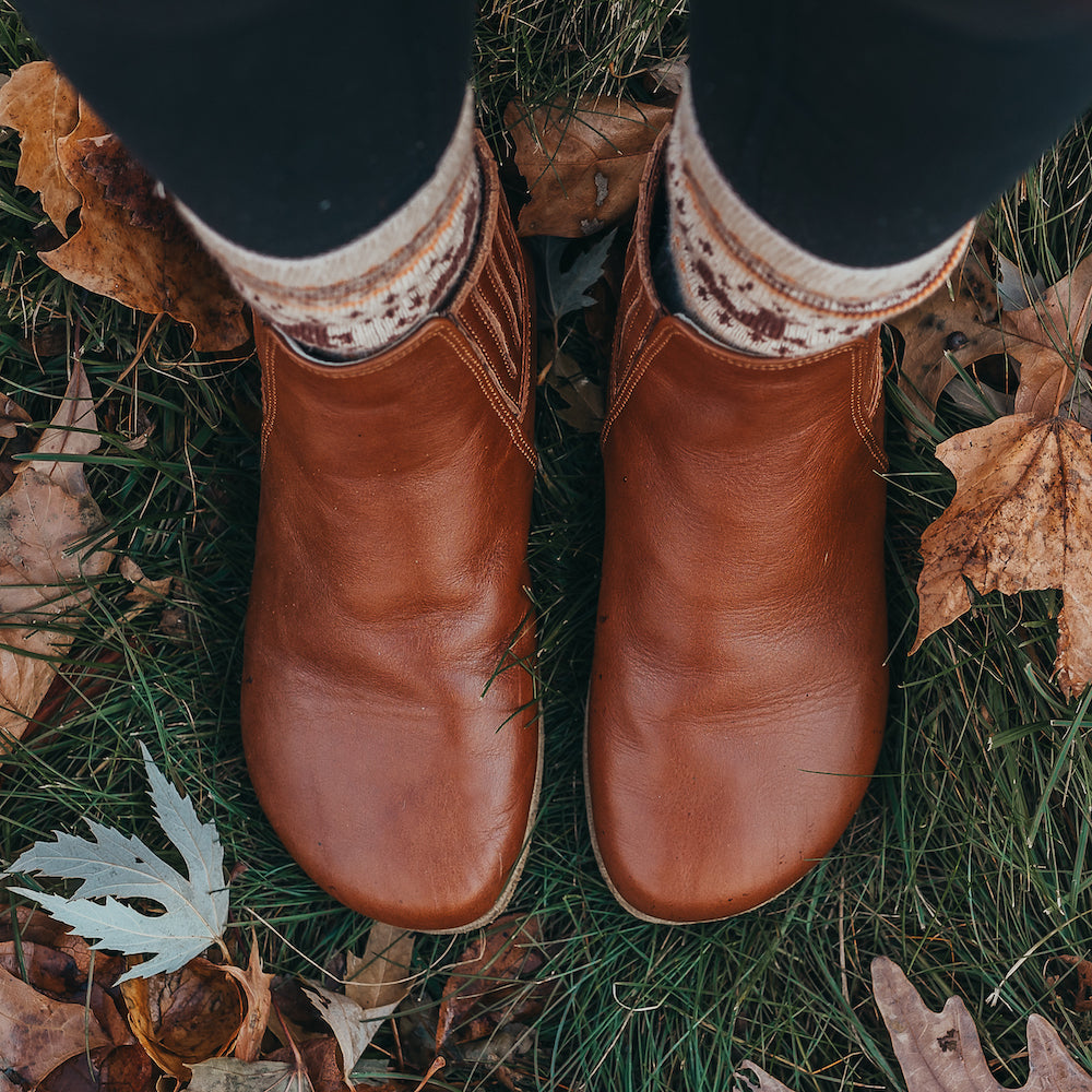 A photo of designed by Anya Lila boots made from leather and rubber soles. The boots are black in color, they are a Chelsea boot style with a lined detailed panel on the sides.  Photo shoes a top down view of both shoes on a person standing in the grass. #color_brown