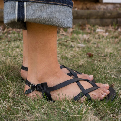 A black leather strappy sandal with a silver buckle in the back. Both sandals are shown facing right on a tan-skinned woman wearing rolled, loose, dark wash jeans standing in grass. #color_black
