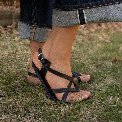 A black leather strappy sandal with a silver buckle in the back. Both sandals are shown facing right on a tan-skinned woman wearing rolled, loose, dark wash jeans standing in grass with her right heel popped. #color_black