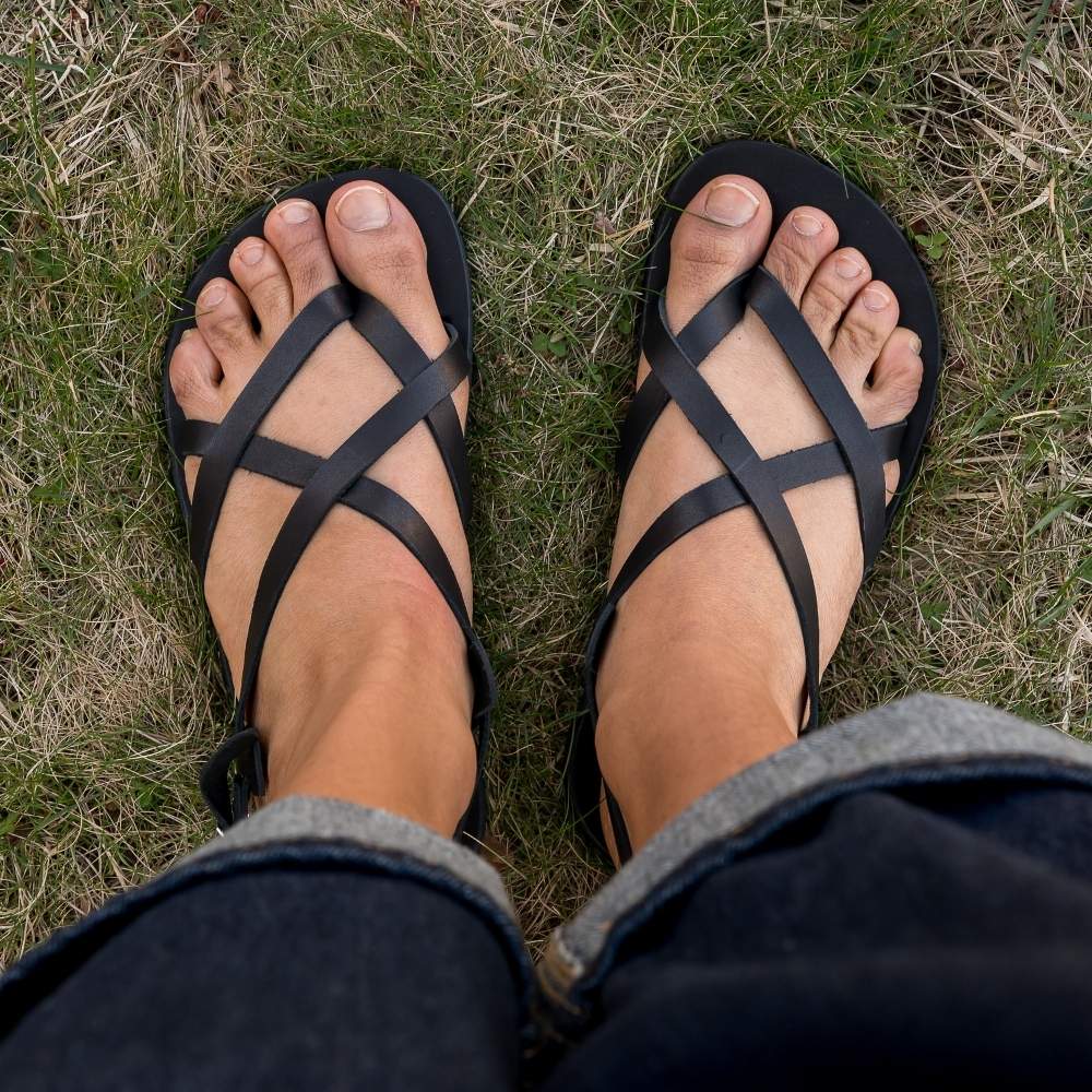A black leather strappy sandal with a silver buckle in the back. Both sandals are shown from above on a tan-skinned woman wearing rolled, loose, dark wash jeans standing in grass. #color_black