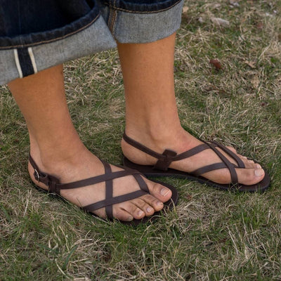 A brown leather strappy sandal with a silver buckle in the back. Both sandals are shown staggered facing right on a tan-skinned woman wearing rolled, loose, dark wash jeans standing in grass. #color_brown