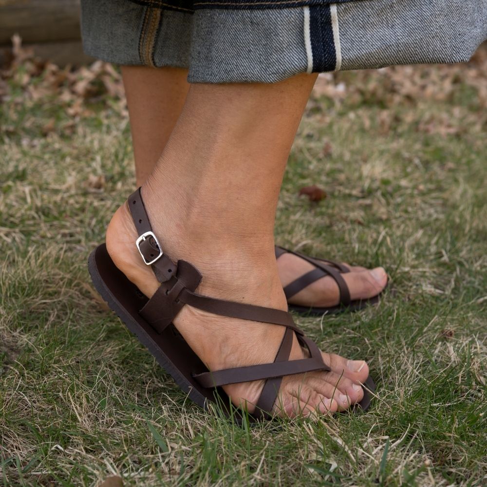 A brown leather strappy sandal with a silver buckle in the back. Both sandals are shown facing right on a tan-skinned woman wearing rolled, loose, dark wash jeans standing in grass with her right heel popped. #color_brown
