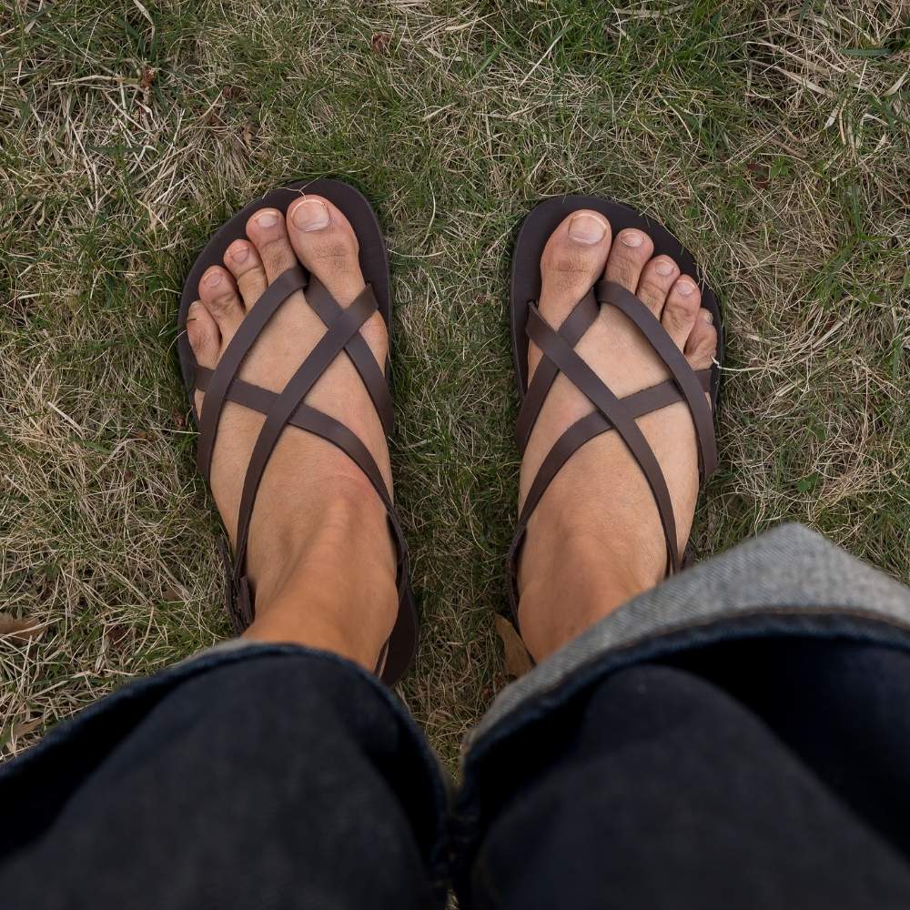 A brown leather strappy sandal with a silver buckle in the back. Both sandals are shown from above on a tan-skinned woman wearing rolled, loose, dark wash jeans standing in grass. #color_brown
