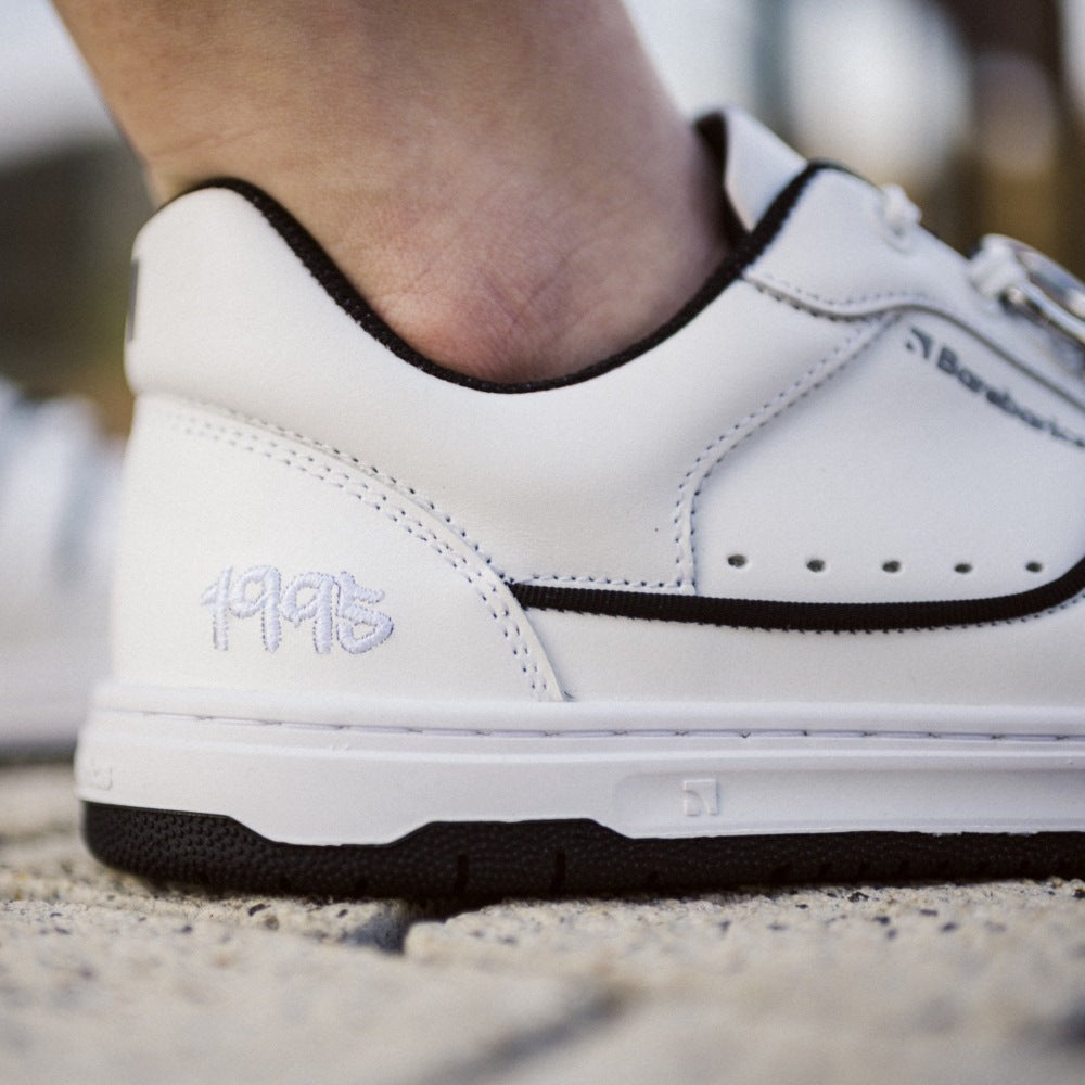 A photo Barebarics Arise chunky leather sneaker in white with black and white soles and white laces. Shoes are perforated on the sides and toe box and have green detailing on the soles, both sides of the shoe, and the lining. Right shoe heel is shown close up here on a person standing on stone pavement. #color_white-black