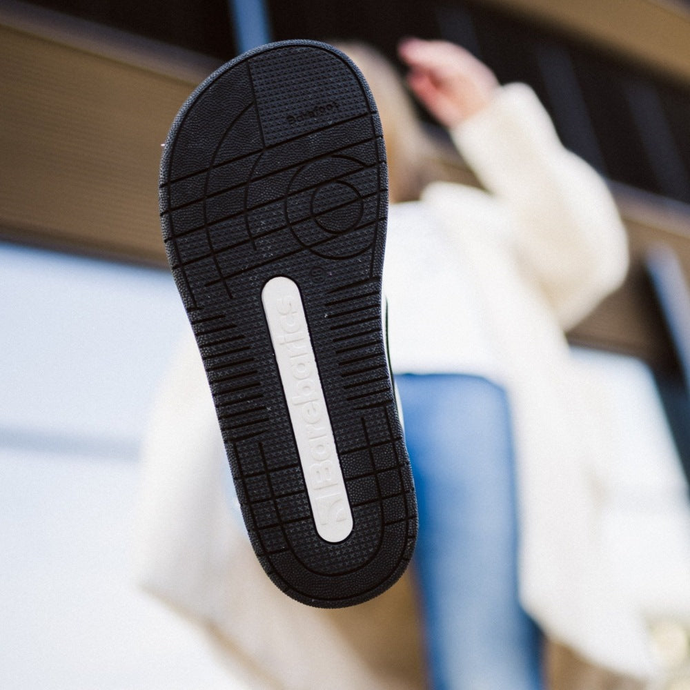 A photo Barebarics Arise chunky leather sneaker in white with black and white soles and white laces. Shoes are perforated on the sides and toe box and have green detailing on the soles, both sides of the shoe, and the lining. Left shoe sole is shown here on a person lifting their leg with a building in the background. #color_white-black