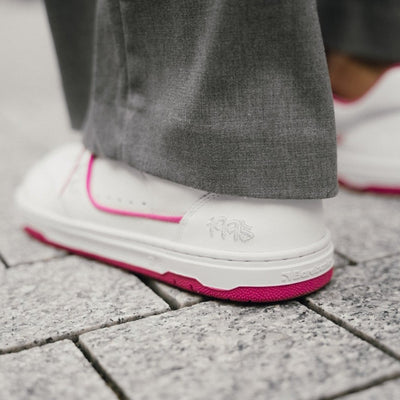 A photo Barebarics Arise chunky leather sneaker in white with pink and white soles and white laces. Shoes are perforated on the sides and toe box and have pink detailing on the soles, both sides of the shoe, and the lining. Left shoe heel is shown close up on a person wearing wide-legged grey dress pants standing on stone pavement. #color_white-raspberry-pink