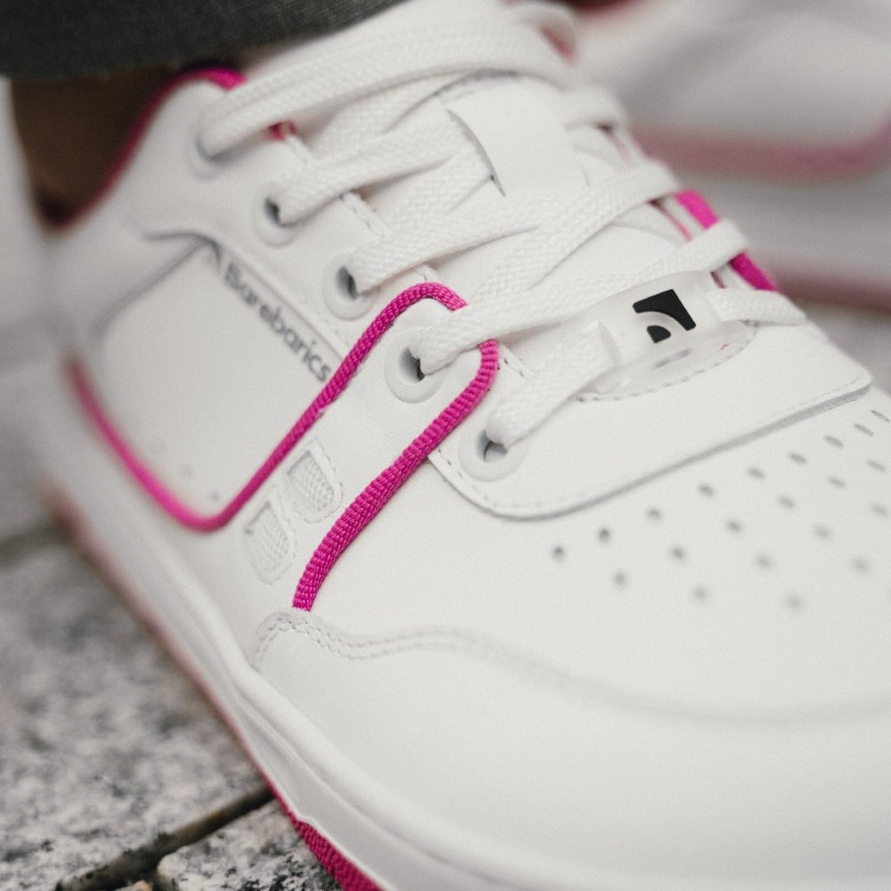 A photo Barebarics Arise chunky leather sneaker in white with pink and white soles and white laces. Shoes are perforated on the sides and toe box and have pink detailing on the soles, both sides of the shoe, and the lining. Left shoe laces are shown close up on a person wearing wide-legged grey dress pants standing on stone pavement. #color_white-raspberry-pink