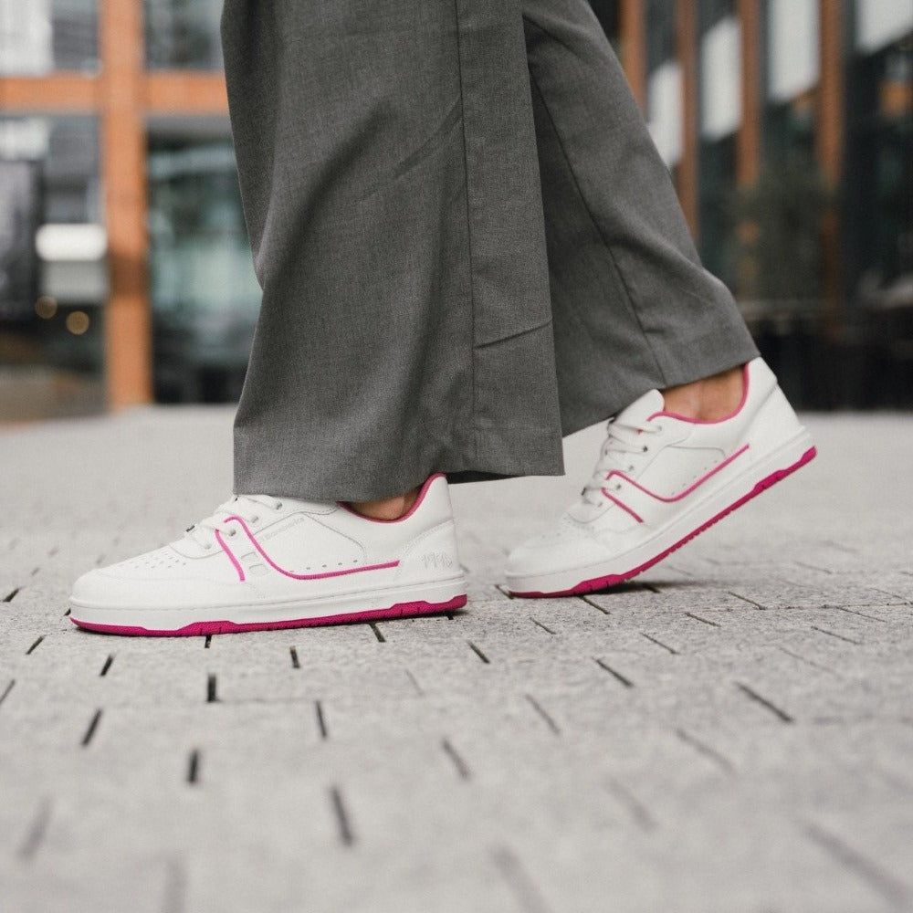 A photo Barebarics Arise chunky leather sneaker in white with pink and white soles and white laces. Shoes are perforated on the sides and toe box and have pink detailing on the soles, both sides of the shoe, and the lining. Both shoes are shown staggered facing left on a person wearing wide-legged grey dress pants standing on stone pavement. #color_white-raspberry-pink