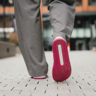 A photo Barebarics Arise chunky leather sneaker in white with pink and white soles and white laces. Shoes are perforated on the sides and toe box and have pink detailing on the soles, both sides of the shoe, and the lining. Both shoes are shown from the back with the right sole lifted and facing the camera on a person wearing wide-legged grey dress pants standing on stone pavement. #color_white-raspberry-pink