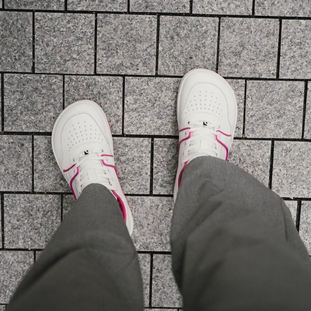 A photo Barebarics Arise chunky leather sneaker in white with pink and white soles and white laces. Shoes are perforated on the sides and toe box and have pink detailing on the soles, both sides of the shoe, and the lining. Both shoes are shown from above on a person wearing grey dress pants standing on a grey brick floor. #color_white-raspberry-pink