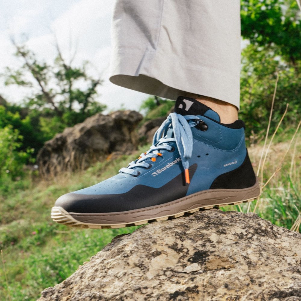 Petrol Blue Barebarics Trekker boot. Shoe is made of blue leather with a black PU toe and heel guard on top. Quick lace hooks are present at the top of the laces. Rubber soles are brown and tan. Left boot is shown facing left on a rock with foliage in the background. Shoe is on a man wearing tan slacks. #color_petrol-blue