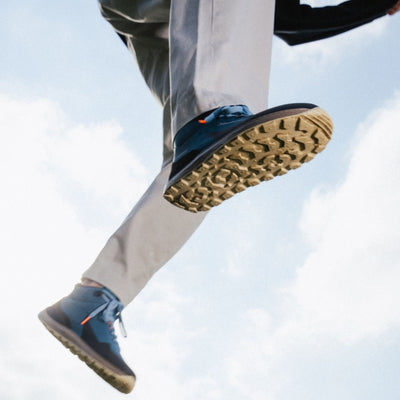 Petrol Blue Barebarics Trekker boot. Shoe is made of blue leather with a black PU toe and heel guard on top. Quick lace hooks are present at the top of the laces. Rubber soles are brown and tan. Both boots are shown staggered from below on a man jumping who is wearing tan slacks. Sky and clouds are in the background. #color_petrol-blue