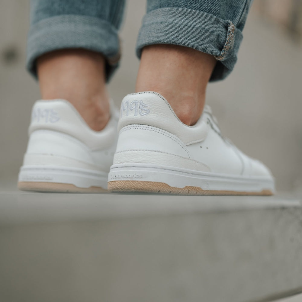 A photo of all white Barebarics Wave classic leath sneakers. Toe box is perforated, the sole is white with tan thread, barebarics brand name is written on the side. Both sneakers are shown here from the back on a woman wearing rolled light denim jeans standing on grey cement wall. #color_all-white