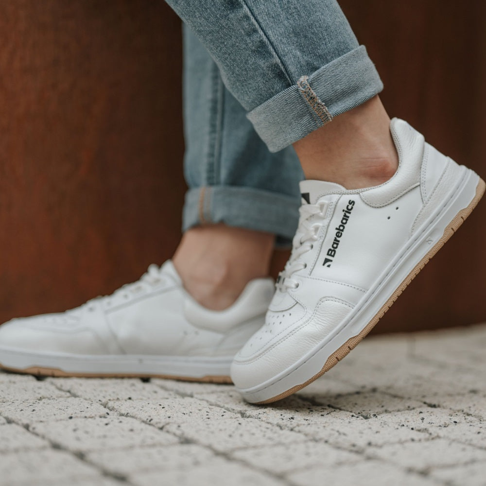 A photo of all white Barebarics Wave classic leath sneakers. Toe box is perforated, the sole is white with tan thread, barebarics brand name is written on the side. Both sneakers are shown here facing left on a woman wearing rolled light denim jeans standing on grey brick pavement with the left foot propped up on the toe. #color_all-white