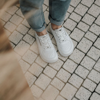A photo of all white Barebarics Wave classic leath sneakers. Toe box is perforated, the sole is white with tan thread, barebarics brand name is written on the side. Both sneakers are shown here from above on a woman wearing rolled light denim jeans standing on grey brick pavement. #color_all-white