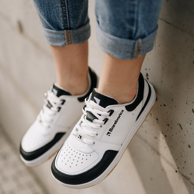 A photo of a black and white classic sneaker with a perforated toe box, the sole is white with tan tread, barebarics brand name is written on the side. Both sneakers are shown diagonally from the top left. Shoes are worn by a woman in rolled light denim jeans sitting on a cement wall. #color_white-black