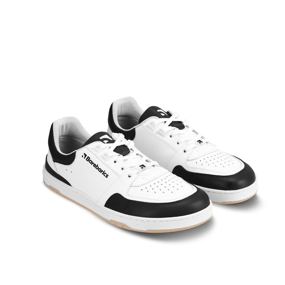 A photo of a black and white classic sneaker with a perforated toe box, the sole is white with tan tread, barebarics brand name is written on the side. Both sneakers are shown from the front angled slightly to the right against a white background. #color_white-black