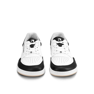 A photo of a black and white classic sneaker with a perforated toe box, the sole is white with tan tread, barebarics brand name is written on the side. Both sneakers are shown beside each other facing towards the front against a white background. #color_white-black