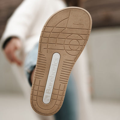 A photo of a black and white classic sneaker with a perforated toe box, the sole is white with tan tread, barebarics brand name is written on the side. Right sneaker sole is shown here. Shoes are worn by a woman in rolled light denim jeans and a long white coat standing with her right foot up to show the sole. #color_white-black