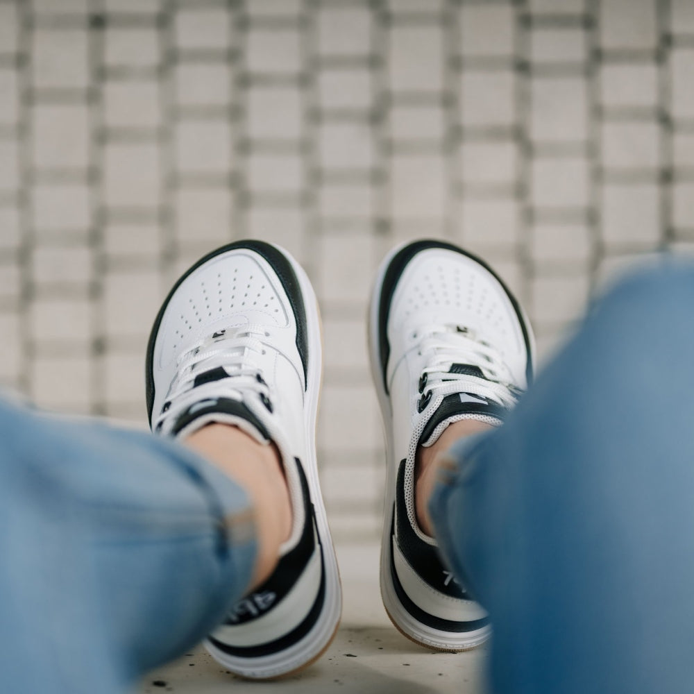 A photo of a black and white classic sneaker with a perforated toe box, the sole is white with tan tread, barebarics brand name is written on the side. Both sneakers are shown from the top down. Shoes are worn by a woman in rolled light denim jeans sitting on a cement wall. #color_white-black
