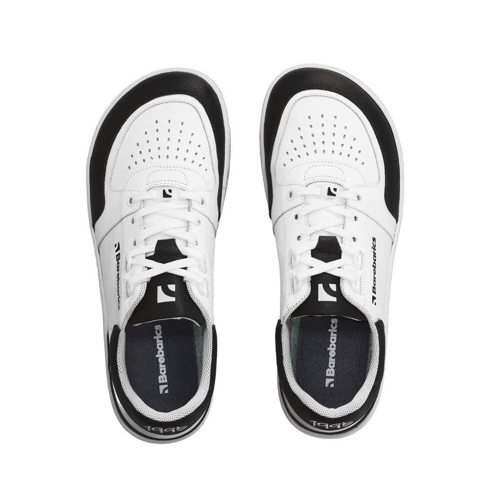 A photo of a black and white classic sneaker with a perforated toe box, the sole is white with tan tread, barebarics brand name is written on the side. Both sneakers are shown from the top down while facing upright against a white background. #color_white-black
