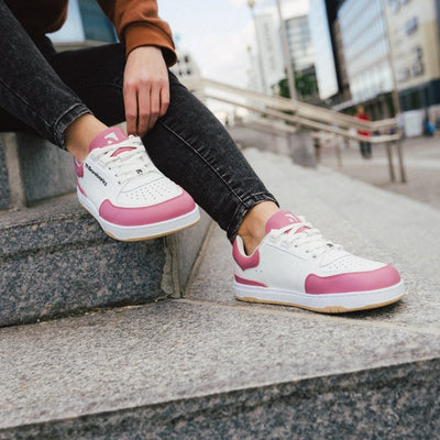 A photo of Barebarics Wave white & bubblegum pink classic sneaker. Shoes are white with a pink toeguard, tongue, heel guard, and at the ankle opening. Toe box is perforated and the sole is white with tan tread. Barebarics brand name is written on the side. Both shoes are shown here facing diagonally right on a woman sitting on cement stairs wearing black skinny jeans and a burnt orange sweatshirt. An urban setting is in the background. #color_white-bubblegum-pink