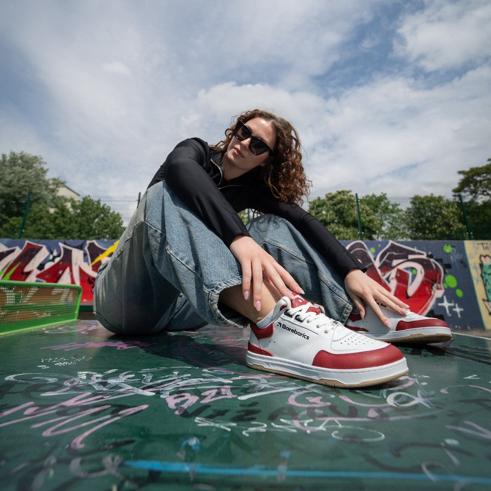 White & Crimson Red Barebarics Wave classic leather sneaker. Shoes are white with a perforated toe box, and a red toe guard, heel guard, ankle guard, and tongue. The sole is white with tan tread, barebarics brand name is written on the side. Both shoes are shown on a woman with curly brown hair. She is wearing wide-legged jeans, a black, long-sleeve shirt, and black sunglasses. She is sitting on a graffitti-covered bench with graffitti-covered walls in the background. #color_white-crimson-red