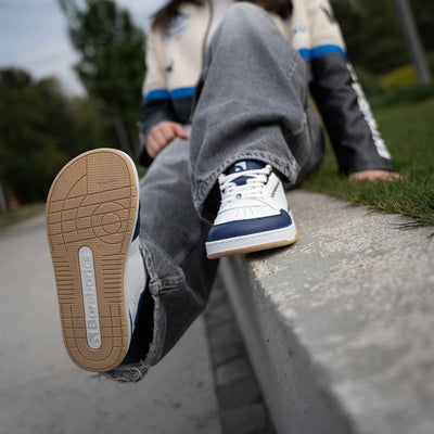 Barebarics Wave white & dark blue classic sneaker. Shoes are white with a dark blue toeguard, tongue, heel guard, and at the ankle opening. Toe box is perforated and the sole is white with tan tread. Barebarics brand name is written on the side. Right shoe is shown here from the front with the left sole shown. Shoes are on a woman sitting on a cement wall wearing baggy grey jeans and a leather jacket that is black on bottom, cream on top, with a blue stripe splitting the two. #color_white-dark-blue