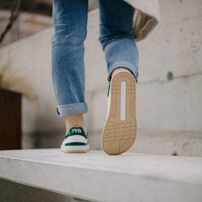 A photo of a green and white classic sneaker with a perforated toe box, the sole is white with tan thread, barebarics brand name is written on the side. Both sneakers are shown from the back with the right shoe propped up showing the sole on a womans wearing rolled light denim blue jeans standing on a cement wall with a cement background. #color_white-dark-green