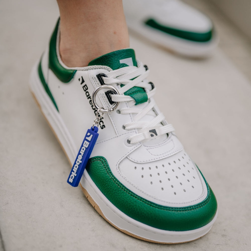 A photo of a green and white classic sneaker with a perforated toe box, the sole is white with tan thread, barebarics brand name is written on the side. Right sneaker is shown diagonally from top right on feet standing on cement. #color_white-dark-green