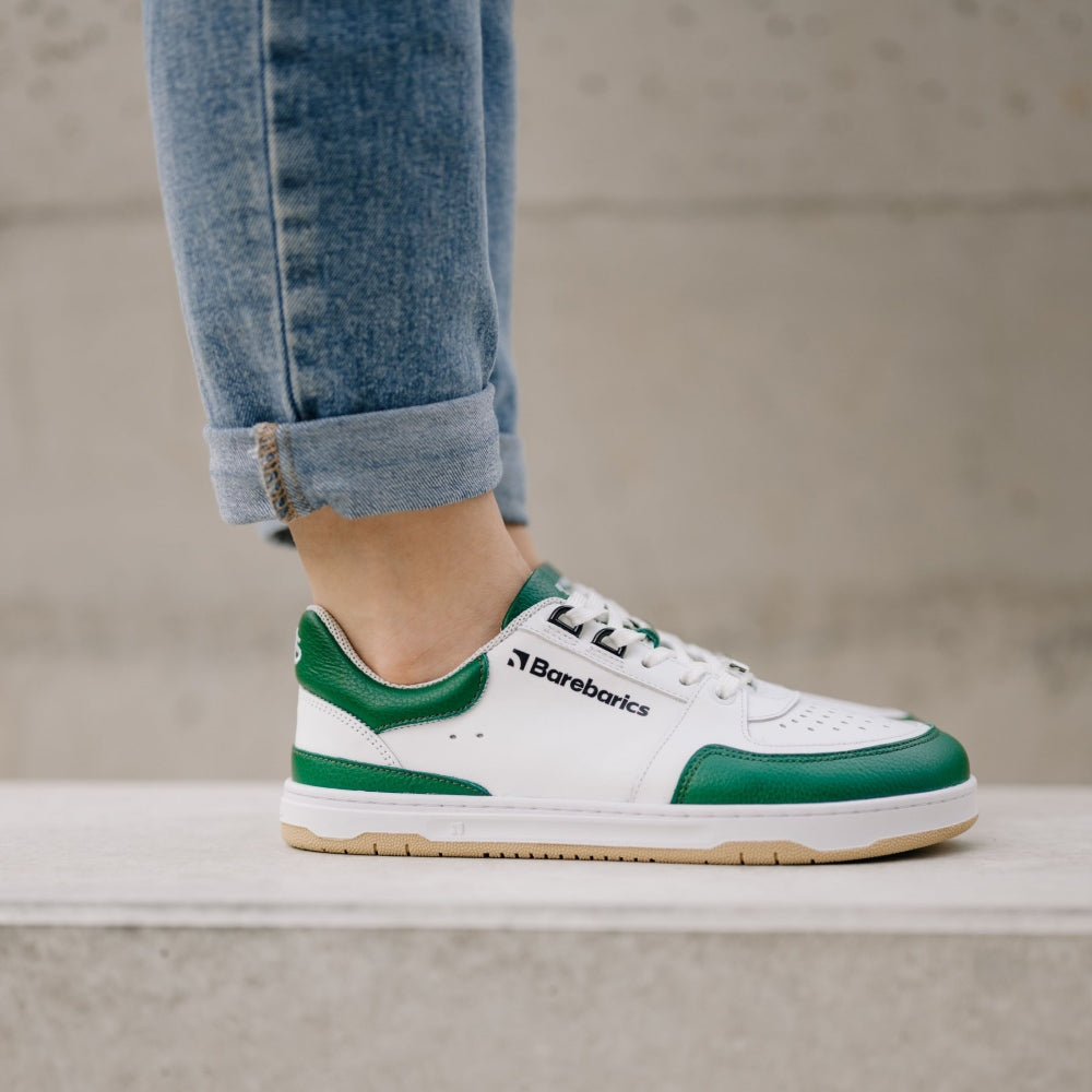 A photo of a green and white classic sneaker with a perforated toe box, the sole is white with tan thread, barebarics brand name is written on the side. Both sneakers are shown from the right side on a womans wearing rolled light denim blue jeans standing on a cement wall with a cement background. #color_white-dark-green