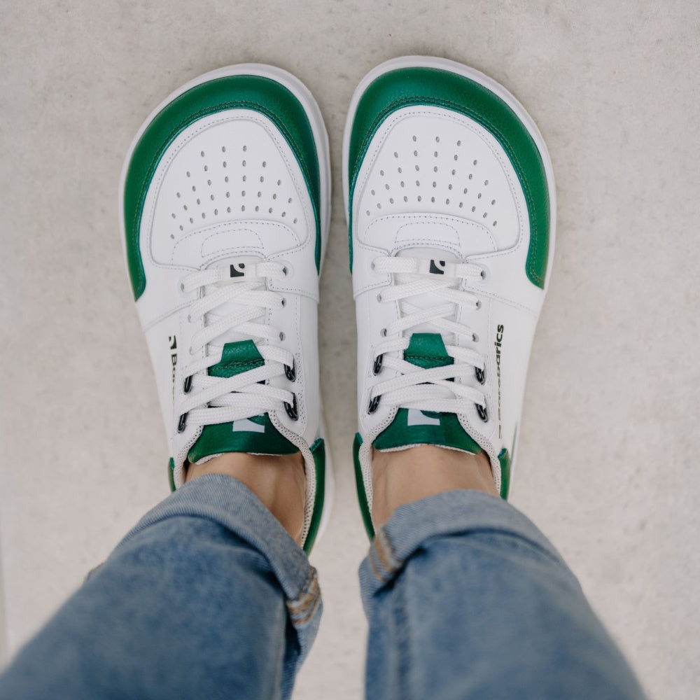 A photo of a green and white classic sneaker with a perforated toe box, the sole is white with tan thread, barebarics brand name is written on the side. Both sneakers are shown from the top down on a womans wearing rolled light denim blue jeans standing on cement. #color_white-dark-green
