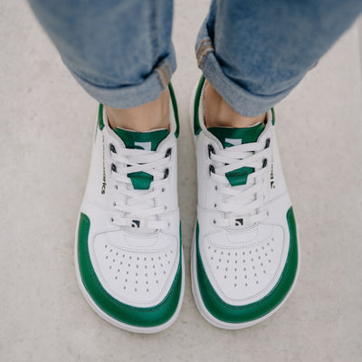 A photo of a green and white classic sneaker with a perforated toe box, the sole is white with tan thread, barebarics brand name is written on the side. Both sneakers are shown from the top down on a womans wearing rolled light denim blue jeans standing on cement. #color_white-dark-green