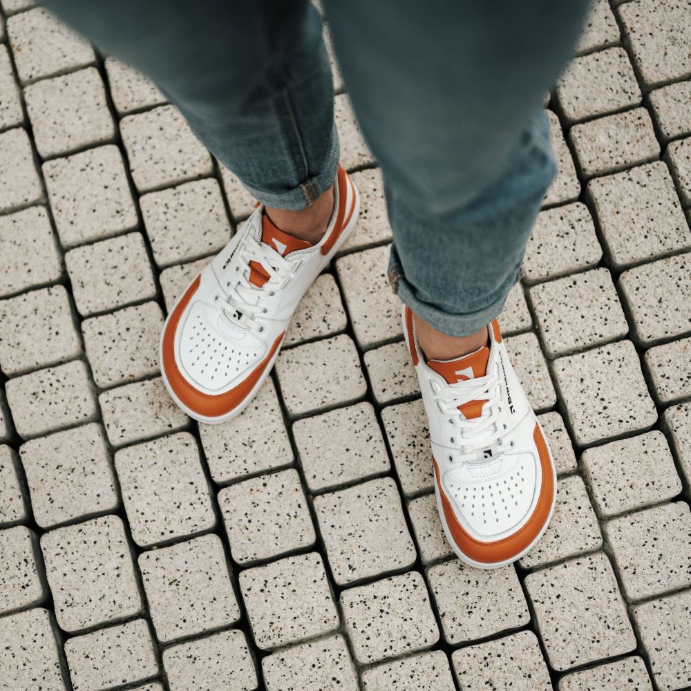 Photo 1 - A photo of Barebarics Wave white & orange classic sneaker with a perforated toe box, the sole is white with tan tread, barebarics brand name is written on the side. Both sneakers are shown here diagonally from the front right against a white background. Photo 2 - Both shoes are shown from the top down on feet against a grey brick background. #color_white-orange
