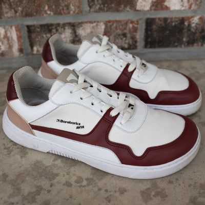 Barebarics Zing sneakers made with a leather upper and a rubber sole. The sneakers are white with burgundy accents around the front and side, and a tan accent over the heel. There are perforated spots on the top of the toe box and barebarics branding on the tongue and side. Both shoes are shown together from the right, on a cement floor in front of a brick wall. #color_burgundy