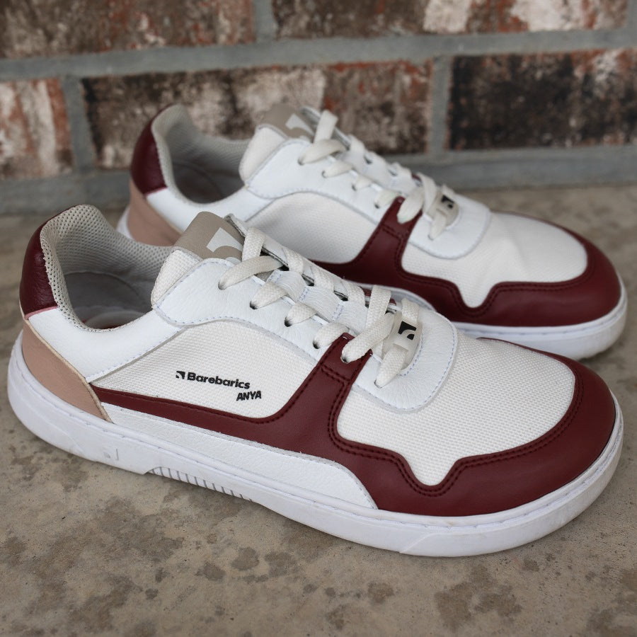 Barebarics Zing sneakers made with a leather upper and a rubber sole. The sneakers are white with burgundy accents around the front and side, and a tan accent over the heel. There are perforated spots on the top of the toe box and barebarics branding on the tongue and side. Both shoes are shown together from the right, on a cement floor in front of a brick wall. #color_burgundy-anya-exclusive