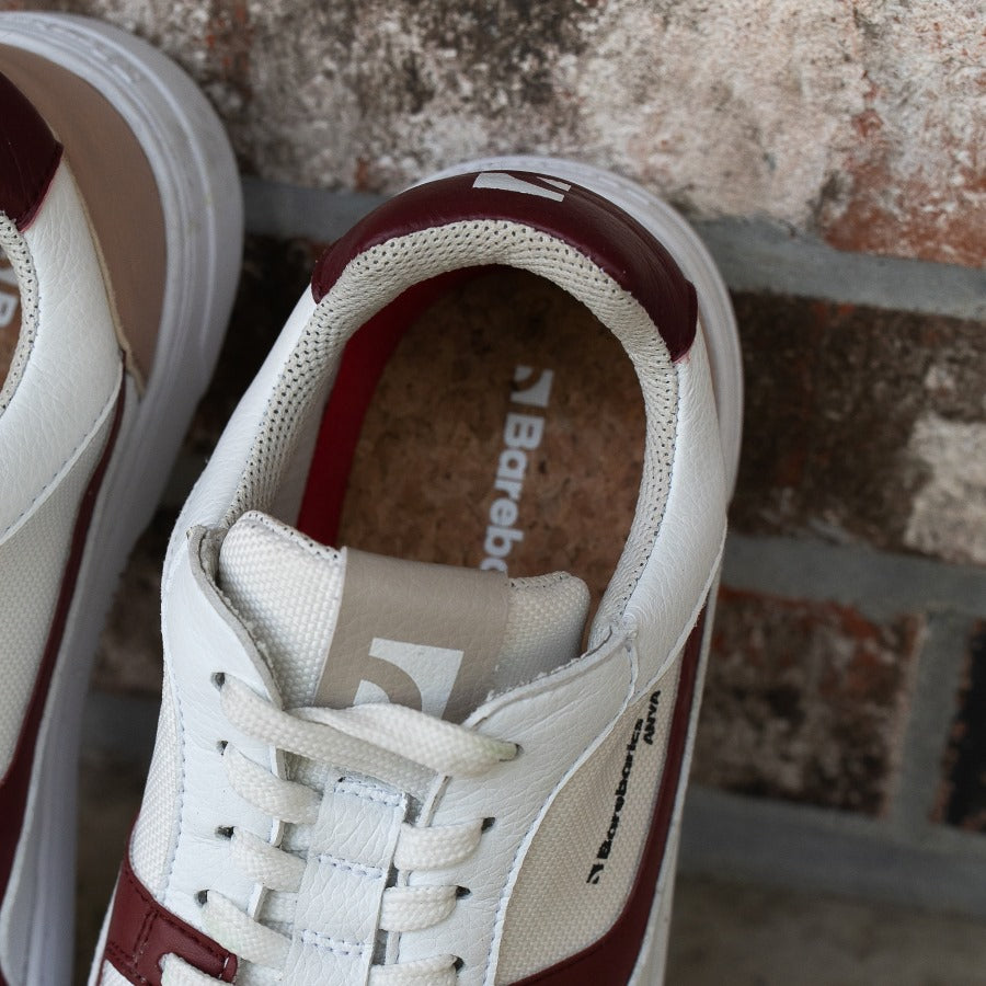 Barebarics Zing sneakers made with a leather upper and a rubber sole. The sneakers are white with burgundy accents around the front and side, and a tan accent over the heel. There are perforated spots on the top of the toe box and barebarics branding on the tongue and side. The heel insole of the left shoe is shown, propped up on a brick wall. #color_burgundy