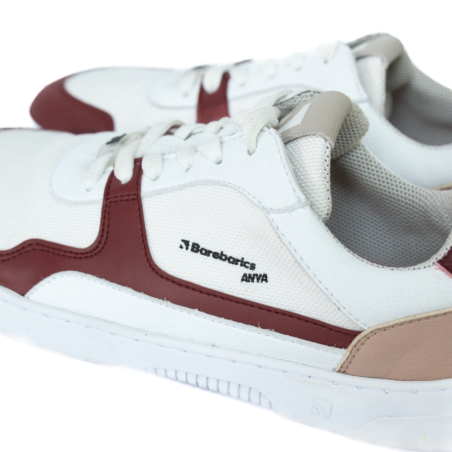 Barebarics Zing sneakers made with a leather upper and a rubber sole. The sneakers are white with burgundy accents around the front and side, and a tan accent over the heel. There are perforated spots on the top of the toe box and barebarics branding on the tongue and side. Both sneakers are shown from the left side with a close up of the Barebarics Anya branding, on a white background. #color_burgundy-anya-exclusive